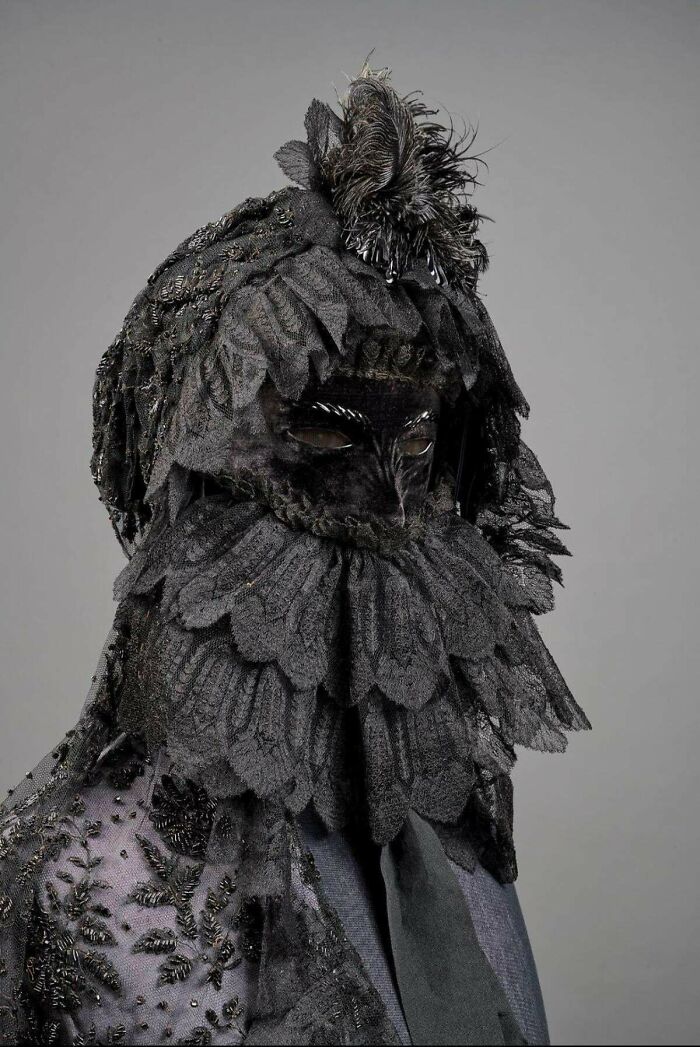 Victorian era costume with intricate lace and feather details, resembling an elaborate black mask.