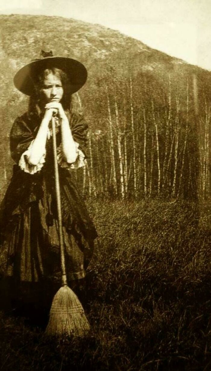 Victorian era woman in field, wearing a hat and holding a broom, with trees and hills in the background.