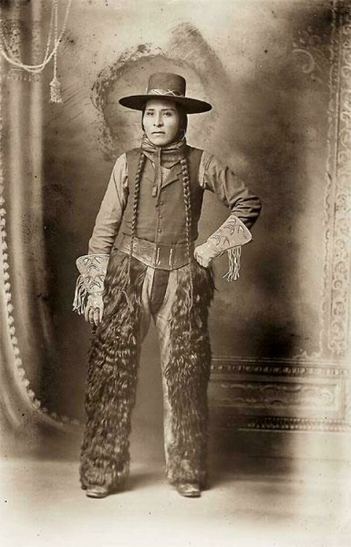 Victorian era cowboy with braids and a wide-brimmed hat, wearing fur chaps and decorated gloves in a vintage studio portrait.