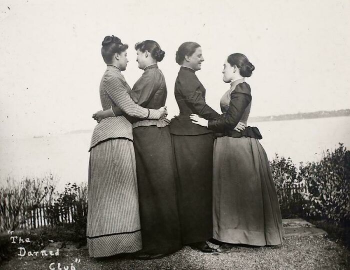 Victorian era women standing in a line outdoors, showcasing period fashion with a scenic lake in the background.