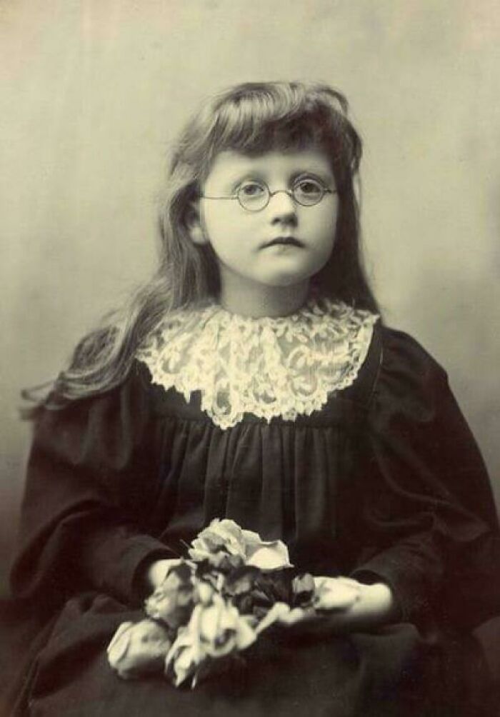 Victorian era girl in glasses and lace collar, holding flowers, showcasing historical fashion.