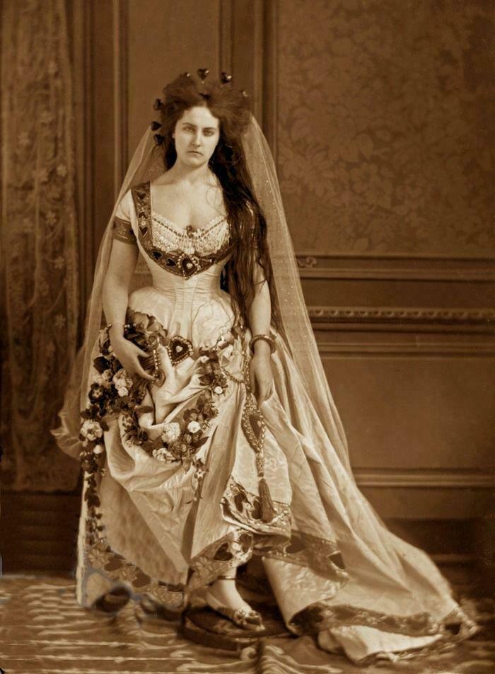 Victorian era woman in elaborate gown adorned with flowers, standing in ornate room.