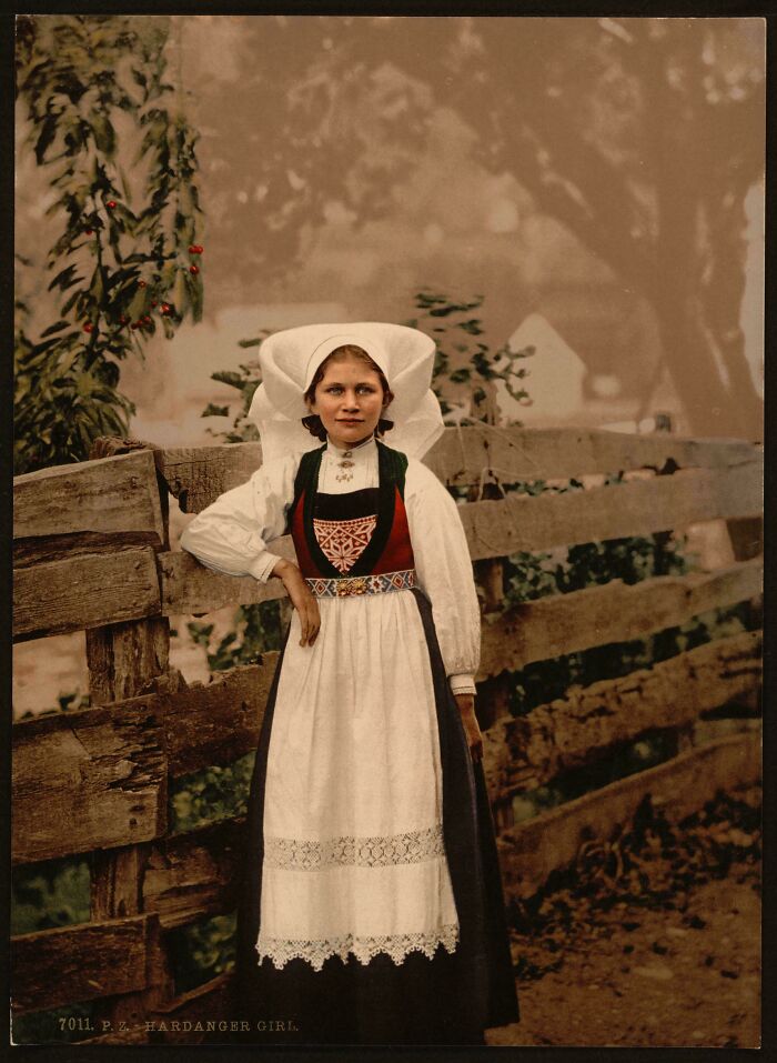 Victorian-era girl wearing traditional attire, standing by a wooden fence with a neutral expression.