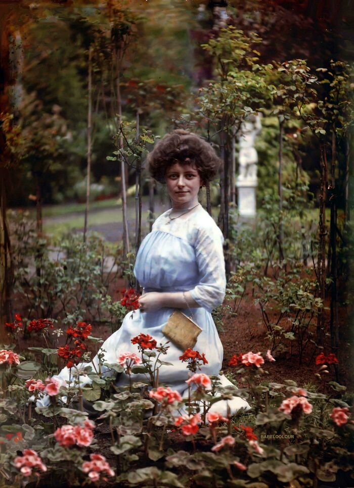 Victorian era woman kneeling in a garden, surrounded by blooming flowers, holding a book.