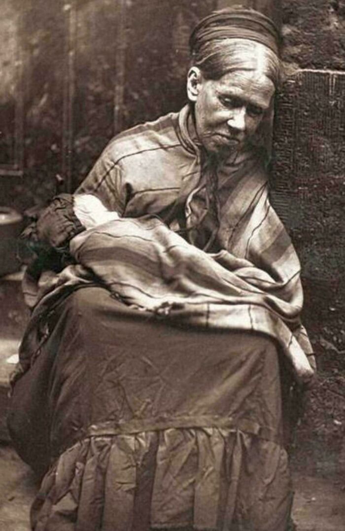 Victorian era woman in period clothing, seated with a solemn expression against a stone wall.