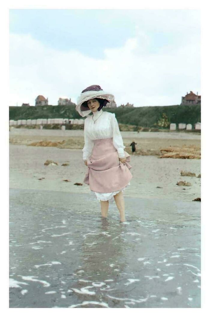 Victorian era woman in a pink skirt and hat wading through shallow water on a beach.
