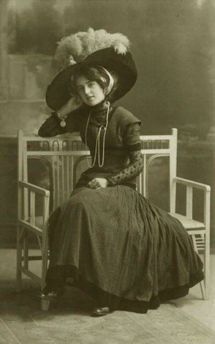 Victorian era woman in elegant dress and large feathered hat seated on a bench.