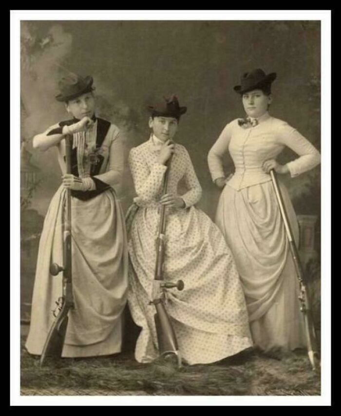Victorian era women posing with rifles, dressed in period attire and hats, showcasing an intriguing historical scene.