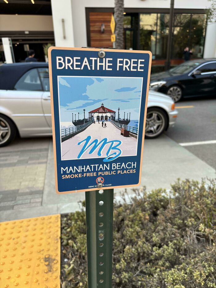 Sign at Manhattan Beach promoting smoke-free public spaces; cars and buildings visible in the background.