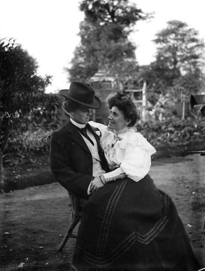 Victorian era couple sitting outdoors, dressed in traditional attire, sharing an intimate moment.