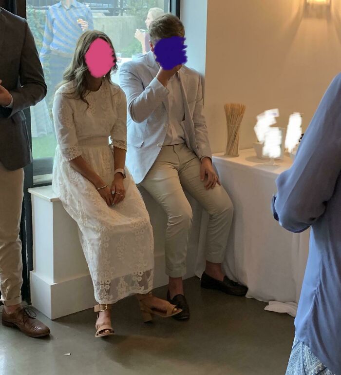 Two wedding guests sit indoors, the woman in a white lace dress, the man in a light suit, both faces obscured.