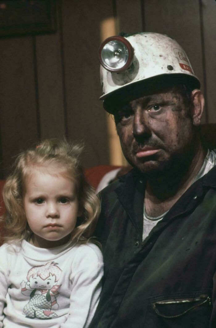 Coal miner with sooty face and hard hat sits beside a young girl, capturing a historical moment unseen in class.
