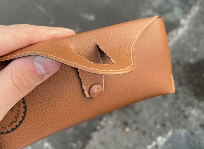 Low-quality item; a hand holds a torn brown leather glasses case, highlighting poor craftsmanship.