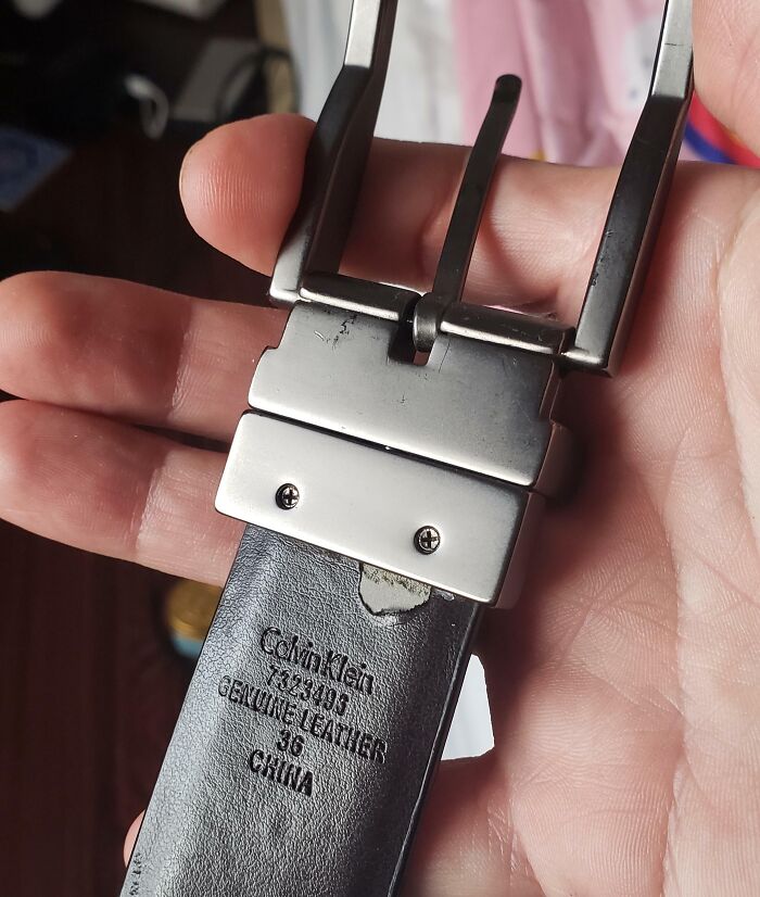 Hand holding a low-quality belt with worn leather and visible screws, labeled "Genuine Leather" and "Made in China."