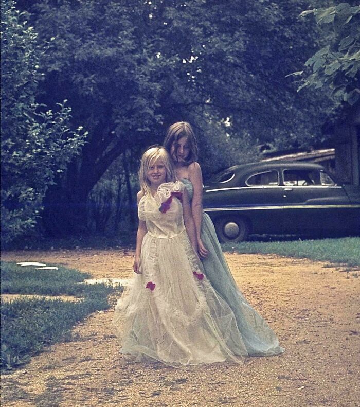 Two young girls in vintage dresses standing on a dirt driveway next to an old car in a historic setting.