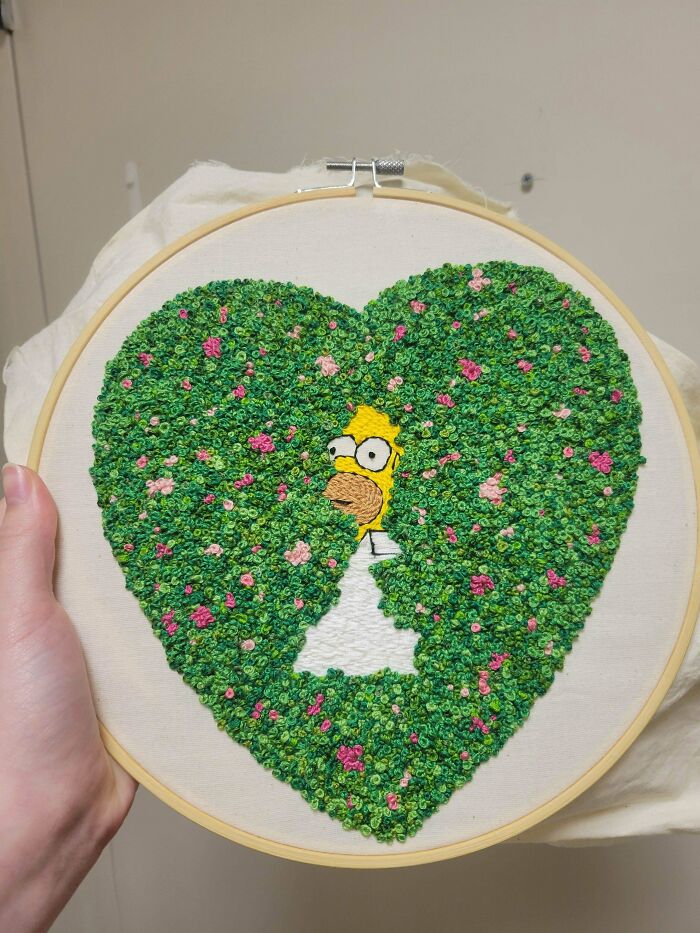 Hand holding embroidery hoop featuring intricate green heart with pink flowers and a hidden yellow character, showcasing embroidery skills.