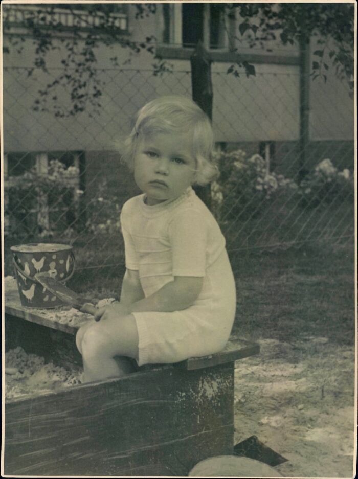Black and white old image of a young child sitting in a sandbox with a bucket and shovel outdoors in a garden.