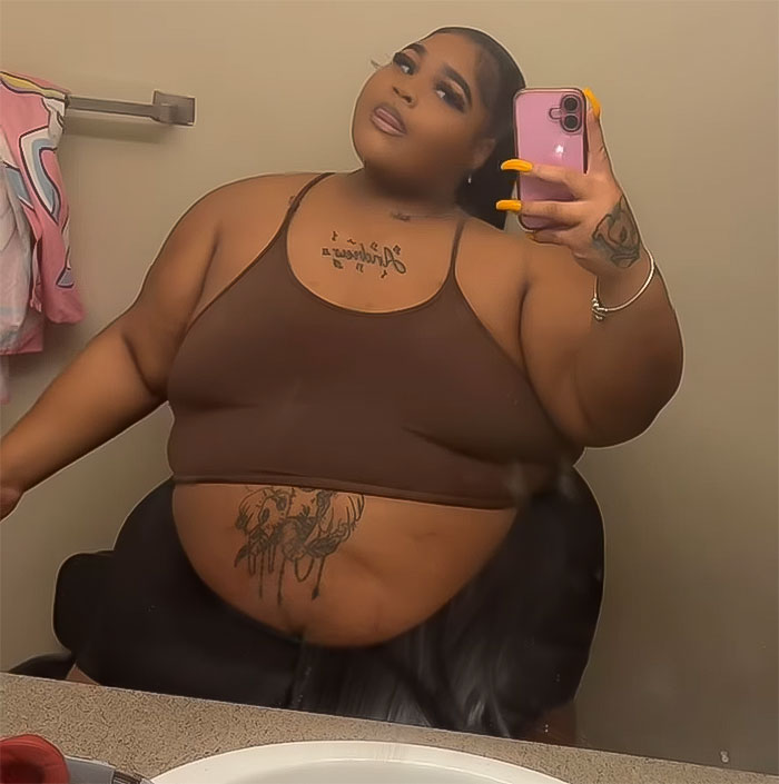 Plus size woman taking a selfie in the mirror, wearing a brown top with tattoos visible.