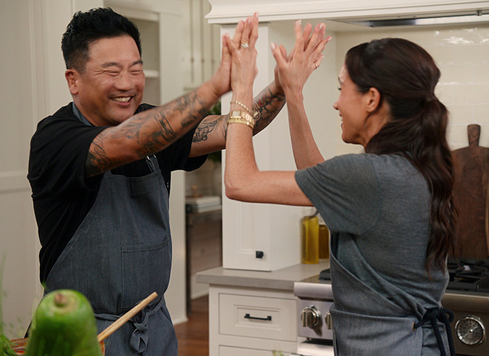 Meghan Markle high-fiving guest in a kitchen, wearing aprons.