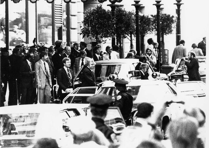 Crowded street scene with police presence during an overlooked historical event, featuring people and vintage cars.