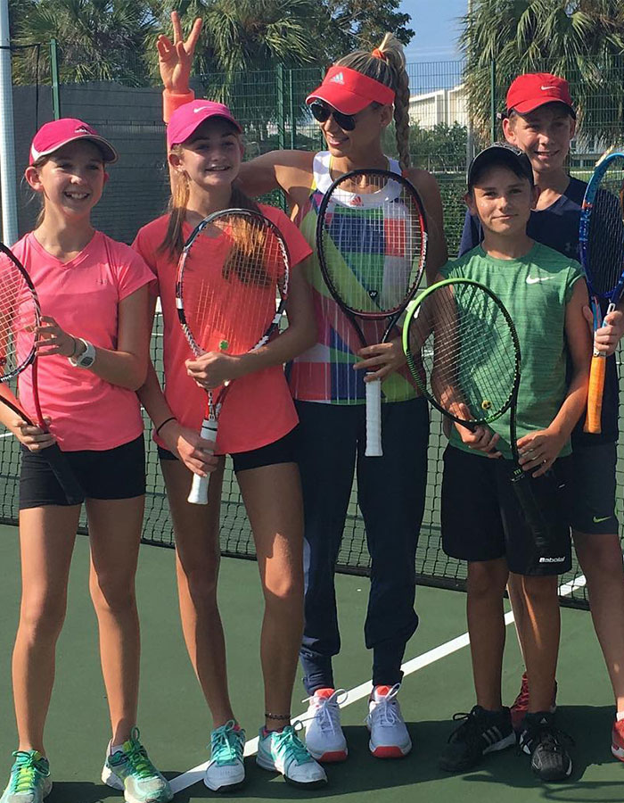 Tennis star with kids on a court, wearing colorful outfit and cap, all holding rackets and smiling.