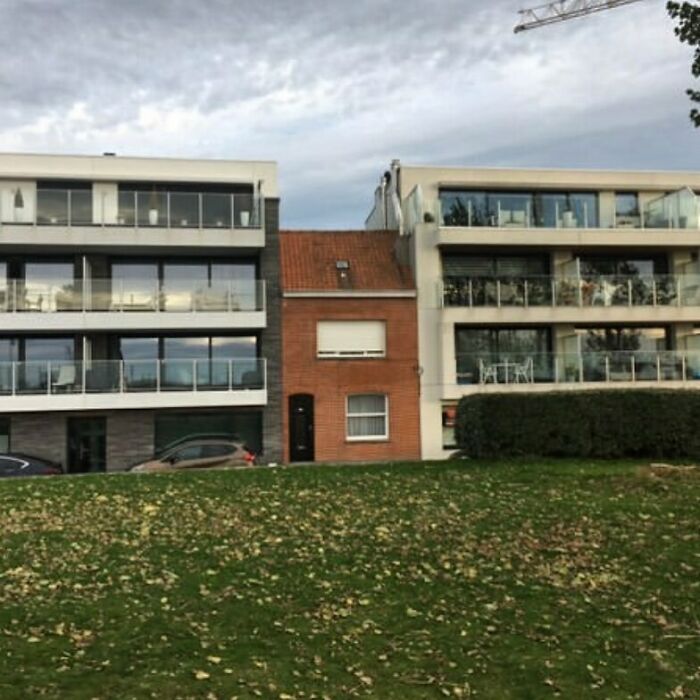 A small brick house squeezed between two modern buildings, exemplifying hilariously bad architectural design.
