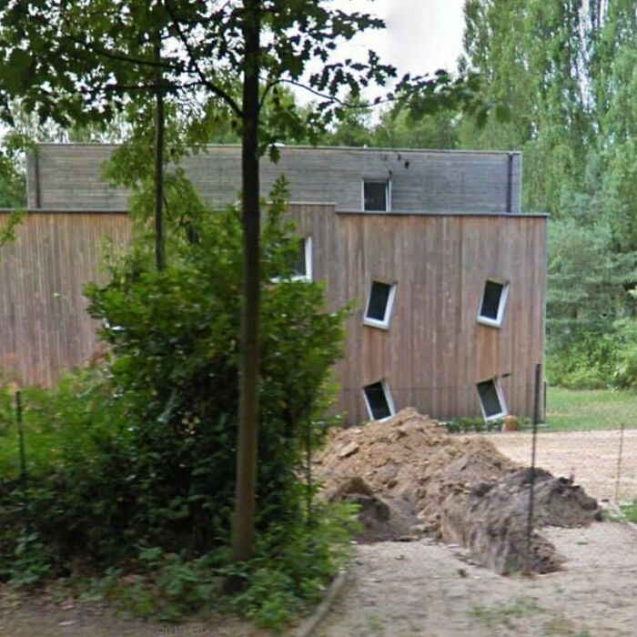 Unusual house with slanted windows and a leaning structure, surrounded by trees and dirt, showcasing hilariously bad architecture.