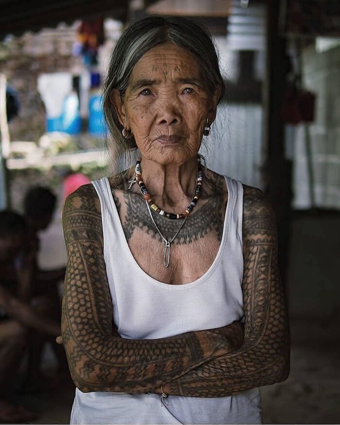 Elderly woman with traditional tattoos on her arms and chest, representing history cool kids, wearing a white tank top.