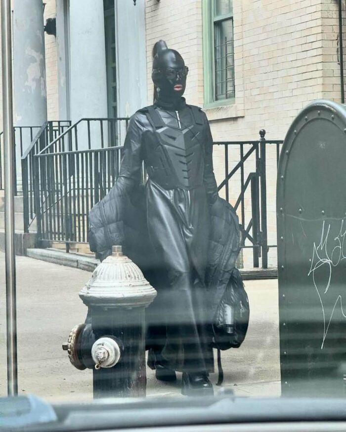 NYC local in experimental fashion, wearing a futuristic black leather outfit and mask, walking down a city street.