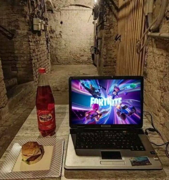 Laptop displaying Fortnite, a burger, and a soda bottle on a wooden table in a dimly lit rustic setting; funny photos theme.