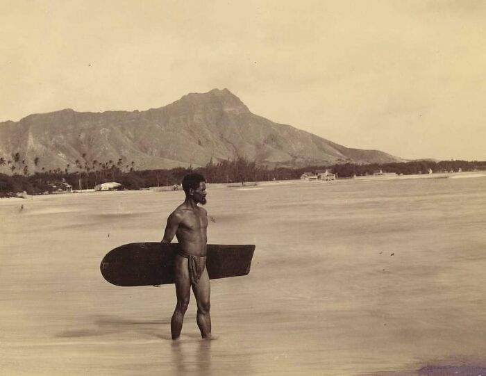 "Man with a vintage surfboard in shallow water, with a mountain in the background, showcasing history cool kids."