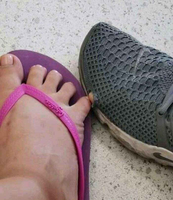 Bizarre photo of a foot in a pink flip-flop pressing against a gray mesh sneaker on a speckled floor.