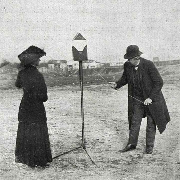 Two people in vintage attire engaging with an old device outdoors, capturing a moment from the past.