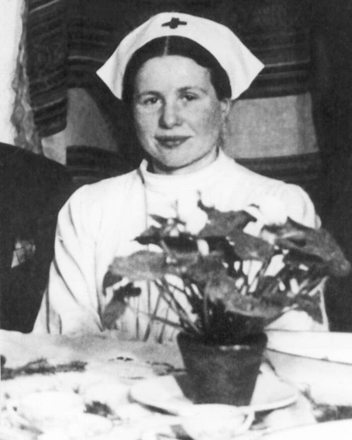 A nurse from history cool kids era, in uniform and cap, sitting at a table with a potted plant in the foreground.