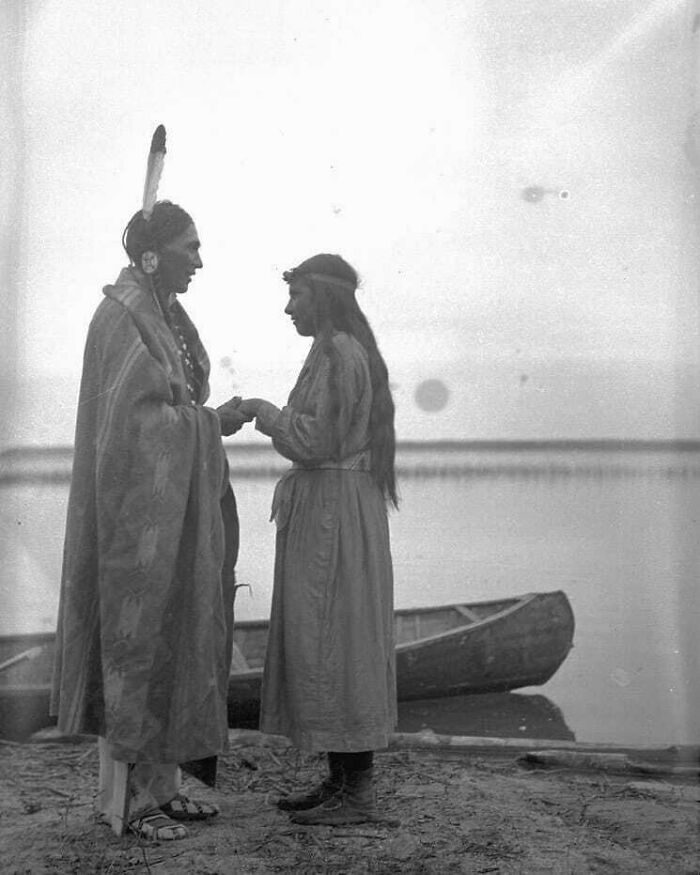 Two people dressed in traditional clothing by a lakeside with a canoe, depicting a moment from history cool kids.