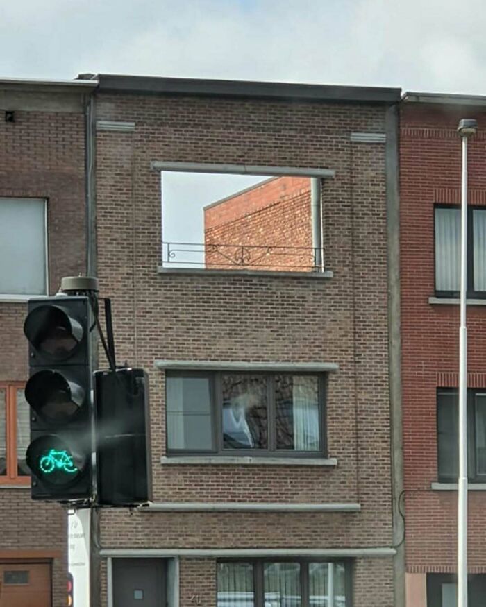 Brick building with a missing window, featuring a railing, next to a bicycle traffic light.