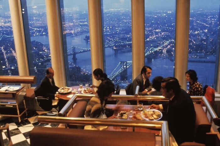 People dining in a high-rise restaurant with a view of a city skyline at night. History Cool Kids vibe.