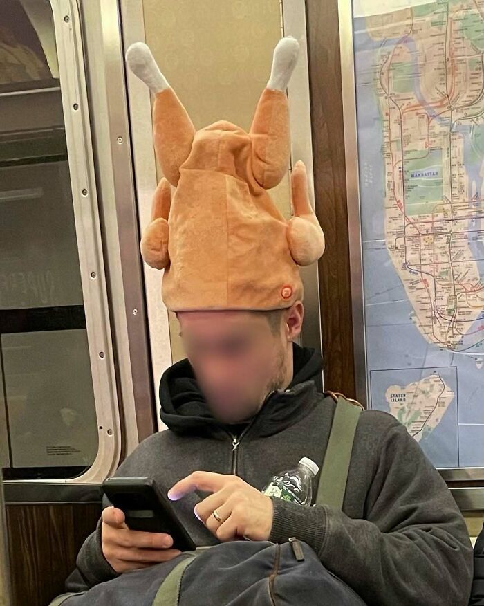 Passenger on subway wearing a funny turkey hat, focused on phone with a subway map in the background.