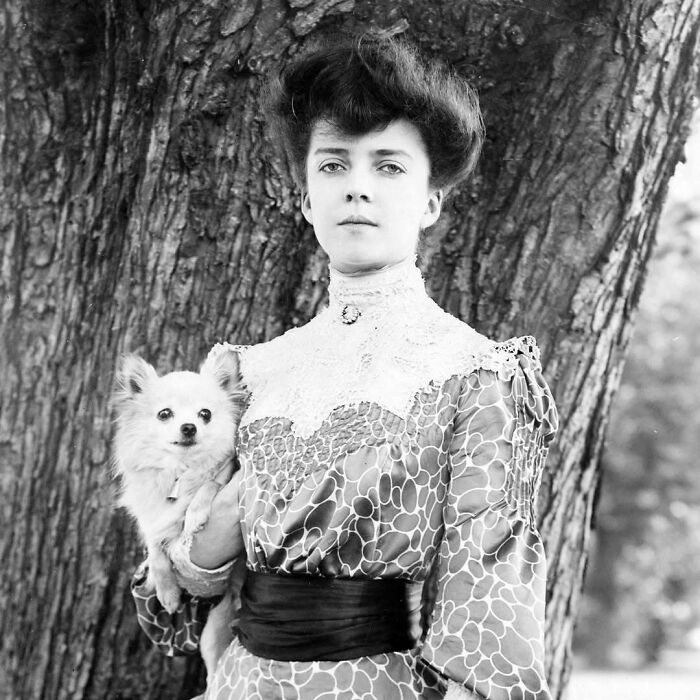 Historical portrait of a woman holding a small dog, standing by a tree, showcasing classic fashion.