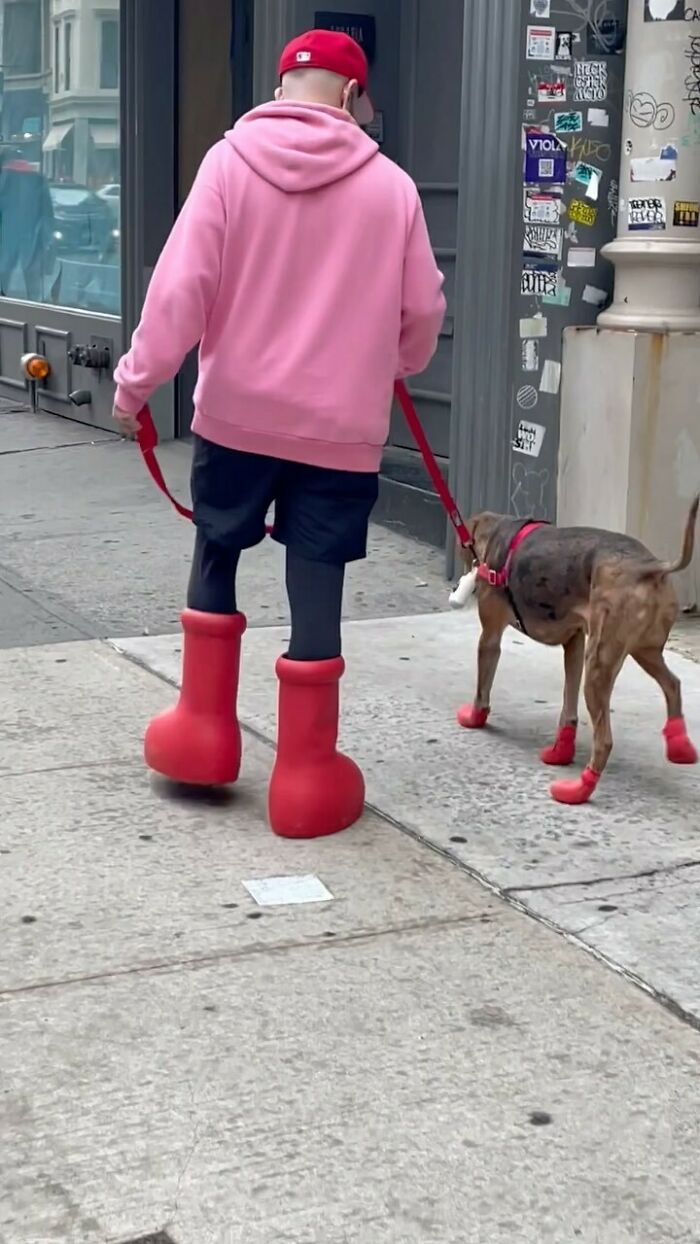 NYC local in pink hoodie and oversized red boots walking a dog in matching red booties, showcasing unique fashion style.