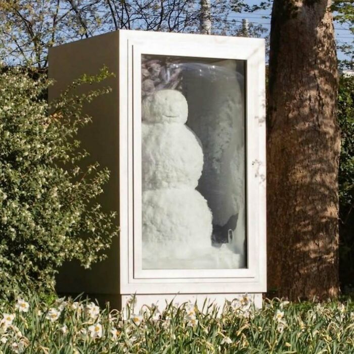Unique garden feature: a snowman preserved in a glass display case amid greenery.
