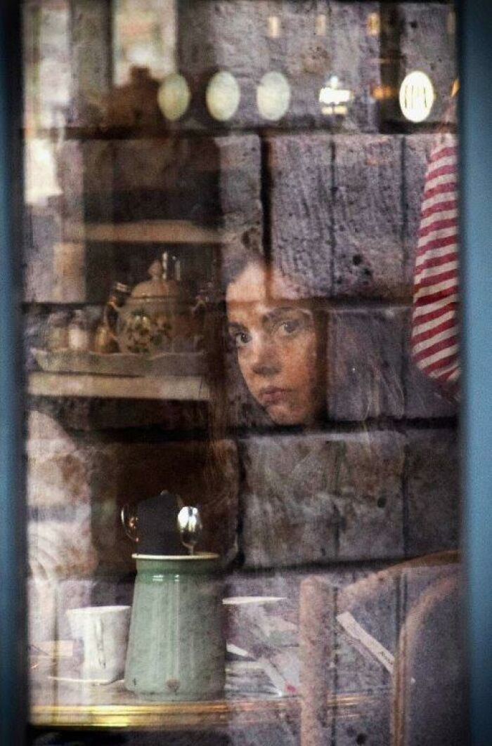 Woman gazing out a window, capturing everyday moments through Nadia Eeckhout's lens, reflections add depth.
