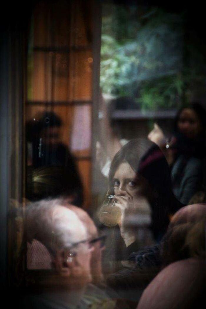 Nadia Eeckhout captures a reflective moment through glass, focusing on a woman sipping a drink in a crowded room.