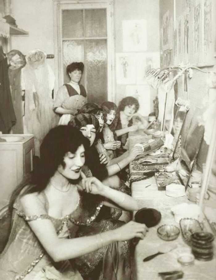 Historical backstage dressing room with women preparing and applying makeup.