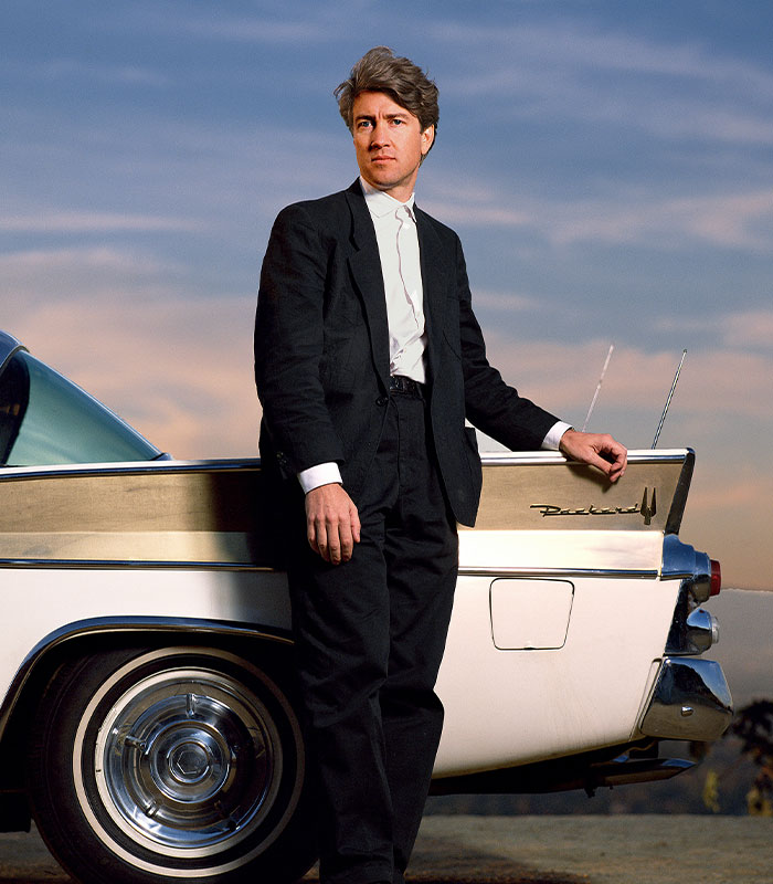 David L***h standing beside a classic car, wearing a black suit, with a serene sunset background.