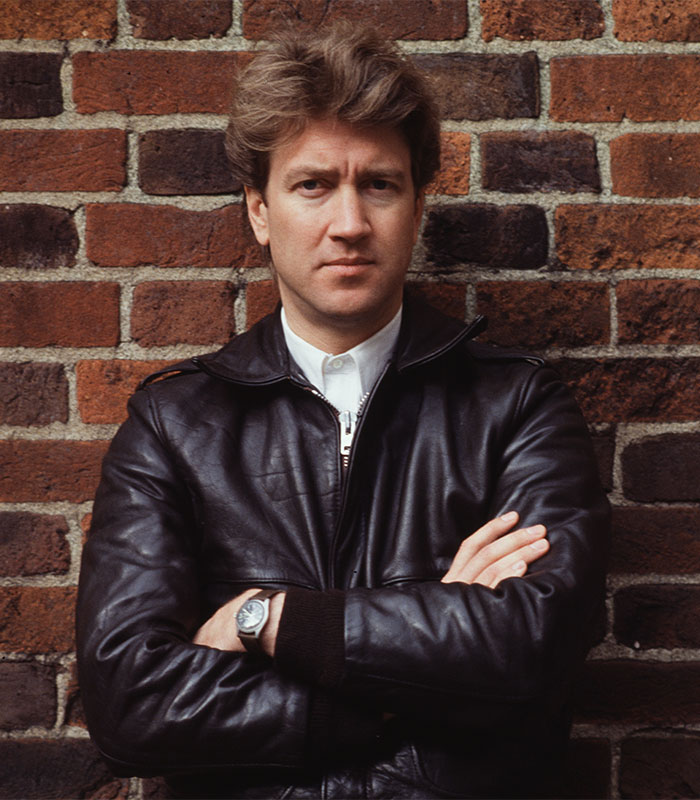 "Man in leather jacket standing against a brick wall, embodying David L***h quotes on questioning reality."
