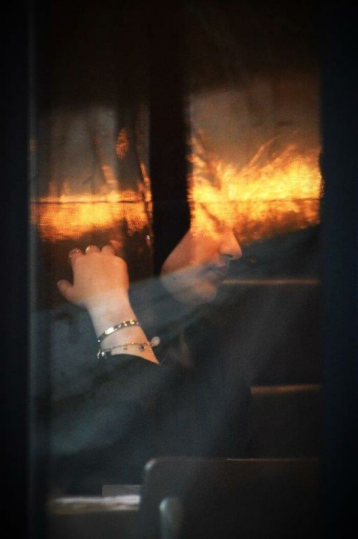 A reflective moment captured through a window by Nadia Eeckhout, featuring a person with bracelets and a soft, warm glow.