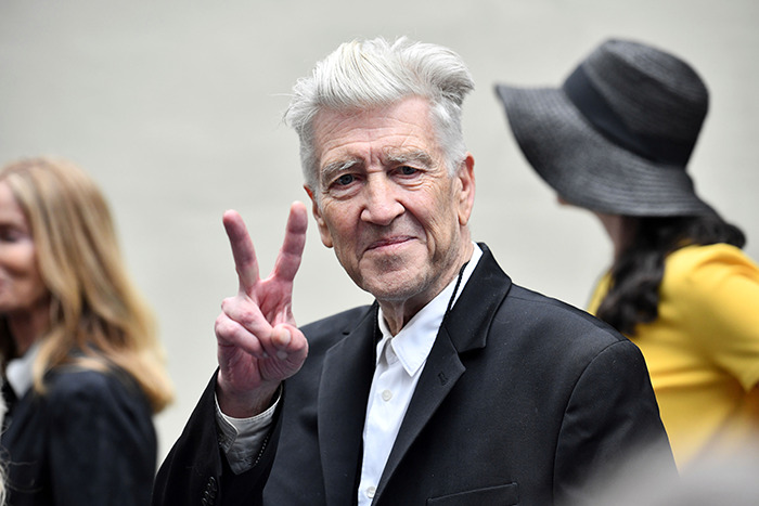David Lynch in a suit with white hair gestures a peace sign, surrounded by people.