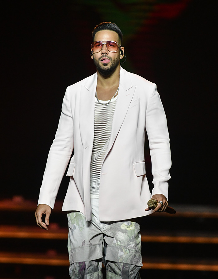 Romeo Santos on stage in a white jacket and floral pants, holding a microphone.