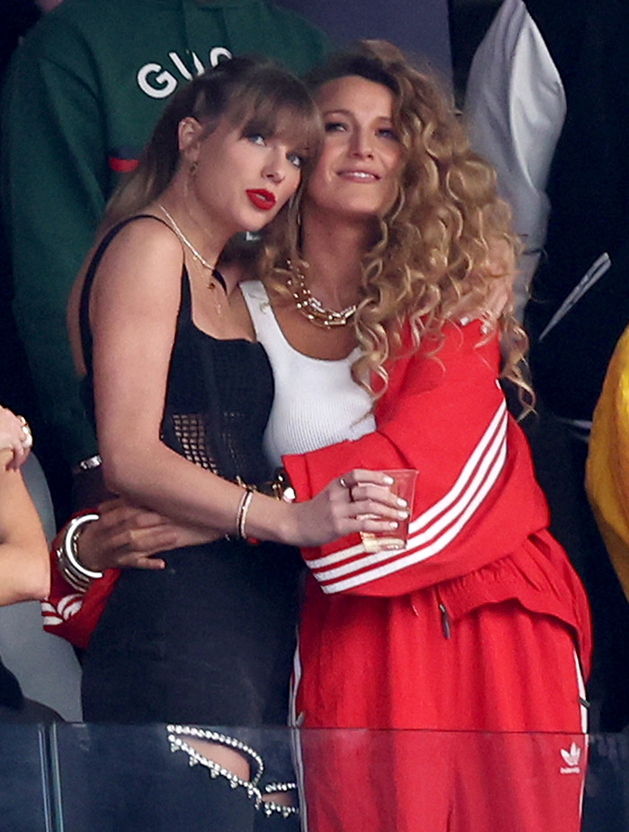 Blake Lively embracing Taylor Swift at an event, one in a black outfit and the other in a red tracksuit.